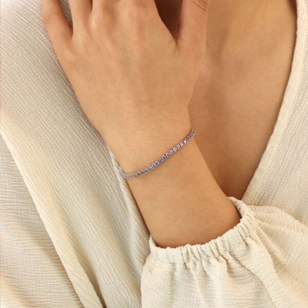 Eine Person, die ein cremefarbenes, strukturiertes, langärmeliges Oberteil trägt, legt ihre Hand auf die Brust und präsentiert das Colour Love Armband Liora Ombre - ein zartes Silberarmband mit kleinen, funkelnden Steinen - an ihrem Handgelenk.