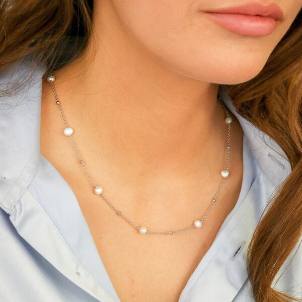 Eine Frau trägt das Baroque Flair Collier Adeline, eine zarte Silberkette mit gleichmäßig verteilten kleinen weißen Perlen, kombiniert mit einem hellblauen Hemd mit Kragen. Das Bild hebt ihren Hals und ihr unteres Gesicht hervor.
