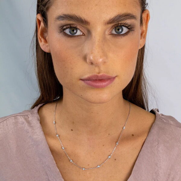 Eine Frau mit glattem braunem Haar und blauen Augen trägt eine hellviolette Bluse und das Tiny Gems Collier Lynette, eine zarte, mit kleinen Edelsteinen besetzte Silberkette, während sie vor einem hellen Hintergrund direkt in die Kamera schaut.
