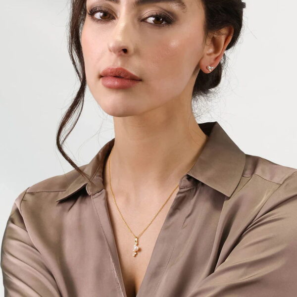 Eine Frau mit gewelltem braunem Haar modelliert das Baroque Flair Collier Linnea - eine goldene Halskette mit Anhänger - zusammen mit einer taupefarbenen Satinbluse und Ohrsteckern und posiert vor einem schlichten hellen Hintergrund.