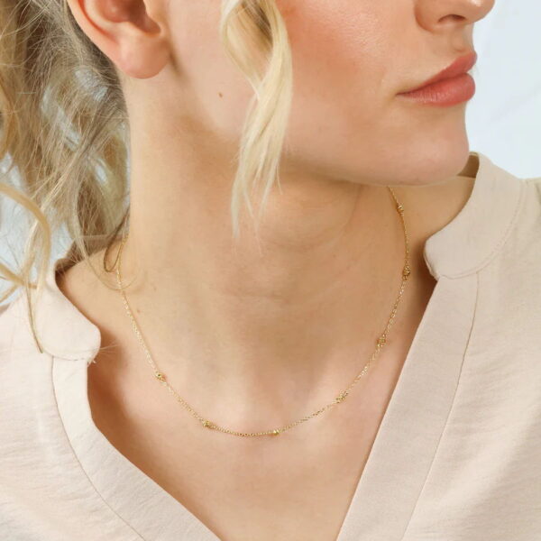 Eine Frau mit heller Haut und blondem Haar trägt das moderne aztekische Collier Orlina, eine zarte goldene Perlenkette, gepaart mit einem cremefarbenen Oberteil mit V-Ausschnitt. Ihr Haar fällt in lockeren Wellen, während sie ihr Gesicht leicht zur Seite dreht.