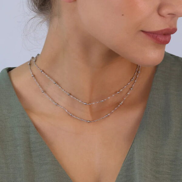 Eine Frau in einem salbeigrünen Oberteil mit V-Ausschnitt trägt das Modern Aztec Collier Darleen, eine zarte zweilagige Silberkette. Die Nahaufnahme hebt ihren Hals, ihr Schlüsselbein und ihr unteres Gesicht hervor.