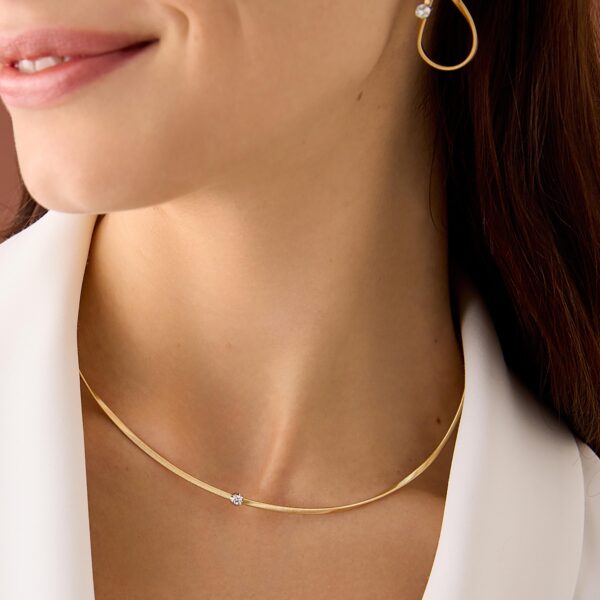 Eine weiß gekleidete Frau trägt das Marco Bicego Marrakech Collier, eine minimalistische Goldkette mit einem kleinen Edelstein und passenden goldenen Ohrringen. Der Fokus liegt auf ihrem Hals und ihrem Schmuck.