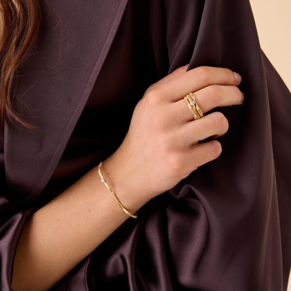 Eine Person in einem dunklen Seidenkleid zeigt ihre Hand, die mit einem goldenen Armband und zwei goldenen Ringen geschmückt ist, darunter der Marco Bicego Marrakech Ring, von denen einer einen kleinen Stein enthält.