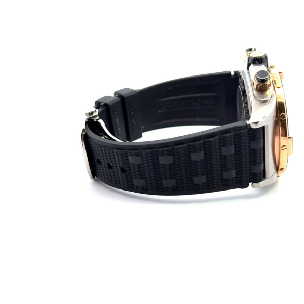 Nahaufnahme der Armbanduhr Breitling Super Chronomat B01 44 mit schwarzem, strukturiertem Kautschukarmband und Metallgehäuse, teilweise geöffnet auf weißem Hintergrund. Der Schriftzug "Breitling" ist auf der Innenseite des Armbands sichtbar.