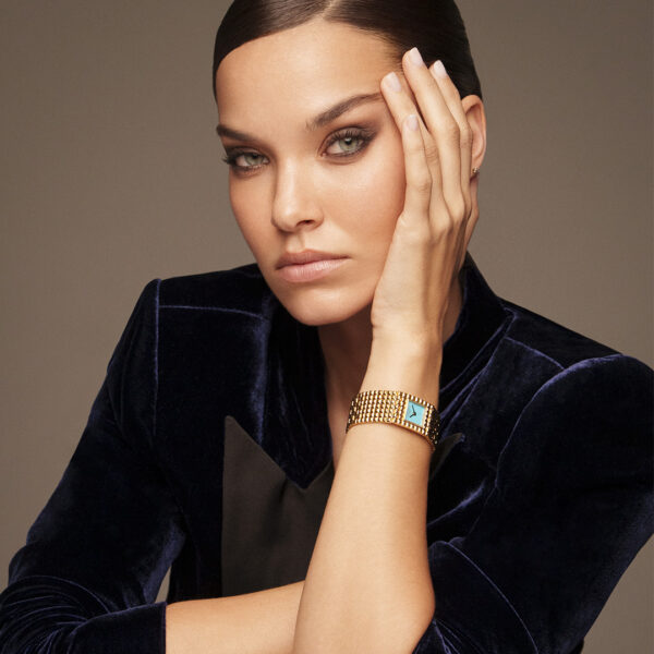 Eine Frau mit hellen Augen und dunklem, zurückgestecktem Haar trägt eine dunkelblaue Samtjacke und das goldene Armband Frederique Constant Classics Manchette mit einem blauen Stein. Sie stützt ihr Gesicht auf eine Hand und blickt mit neutralem Blick in die Kamera.