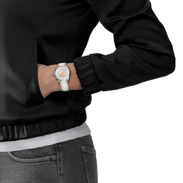 Eine Person in schwarzer Jacke und grauer Jeans steht mit einer Hand in der Tasche und präsentiert die Tissot PR 100 34MM Armbanduhr mit weißem Band und rundem, silber-goldenem Zifferblatt.