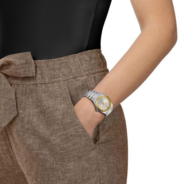 Eine Person in einer hochtaillierten braunen Leinenhose mit gebundenem Gürtel und einem schwarzen Oberteil hat die Hand in der Tasche und präsentiert die Tissot PRX Goldlünette 35MM Armbanduhr.