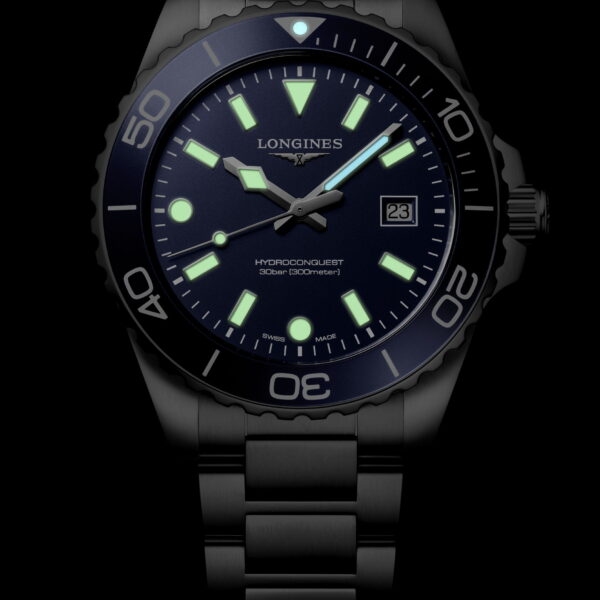Die Longines HydroConquest Quarz verfügt über ein Metallarmband, ein dunkles Zifferblatt mit grün leuchtenden Indexen, Zeigern und Ziffern sowie ein Datumsfenster bei 3 Uhr – alles vor schwarzem Hintergrund.