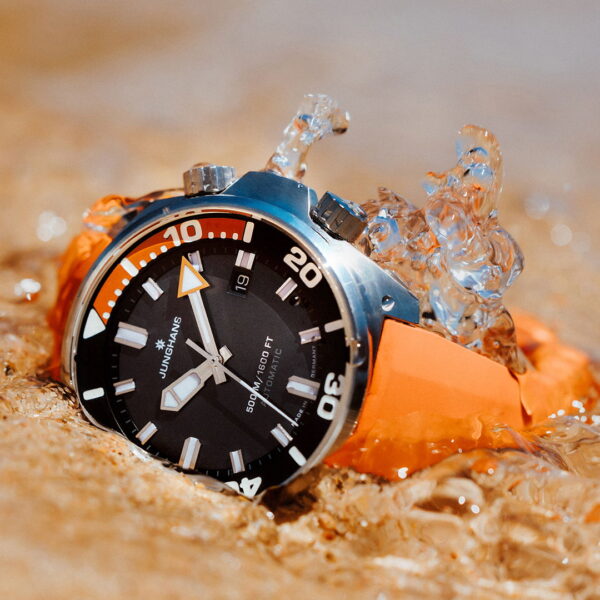 Die Junghans Aquaris Diver mit orangefarbenem Armband und schwarzem Zifferblatt mit Datumsanzeige und auffälligen weißen Markierungen ruht teilweise im flachen Sand und Wasser, während die Spritzer diese robuste Taucheruhr umgeben.