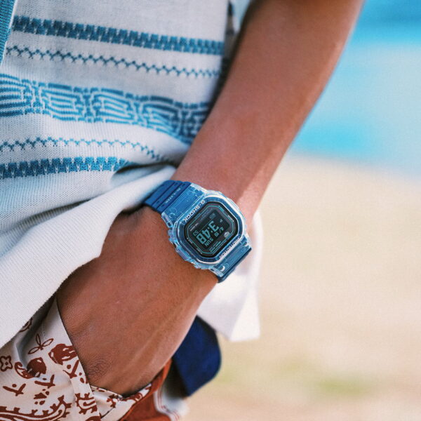 Eine Person in einem weiß-blau gemusterten Hemd und Shorts steht mit einer Hand in der Tasche am Strand und trägt eine Casio G-Shock Pro G-Lide; Meer und Sand verschwimmen sanft im Hintergrund.