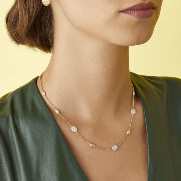 Eine Frau, die das „Marco Bicego Siviglia Collier“ trägt – eine goldene Kette mit runden, strukturierten Perlen –, kombiniert mit einer grünen Bluse; die Aufnahme zeigt sie von den Schultern aufwärts vor einem hellgelben Hintergrund.