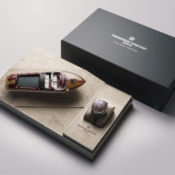 Ein luxuriöses Frederique Constant Classics Runabout Automatic GMT-Geschenkset mit einer eleganten Armbanduhr in einer beigen Vitrine, einem detailgetreuen Yachtmodell aus Holz und einer dunkelgrauen Markenbox.