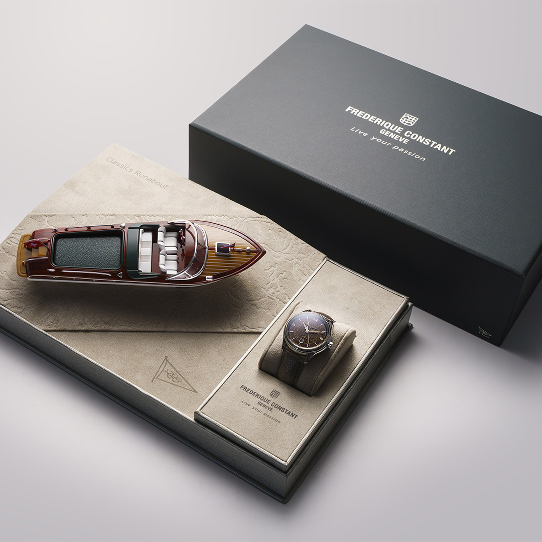 Ein luxuriöses Frederique Constant Classics Runabout Automatic GMT-Geschenkset mit einer eleganten Armbanduhr in einer beigen Vitrine, einem detailgetreuen Yachtmodell aus Holz und einer dunkelgrauen Markenbox.