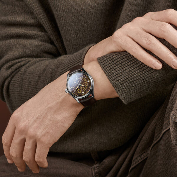 Eine Person in einem braunen Pullover und einer braunen Hose sitzt mit verschränkten Armen da und präsentiert die Armbanduhr „Frederique Constant Classics Runabout Automatic GMT“ mit dunklem, rundem Zifferblatt und braunem Lederarmband.