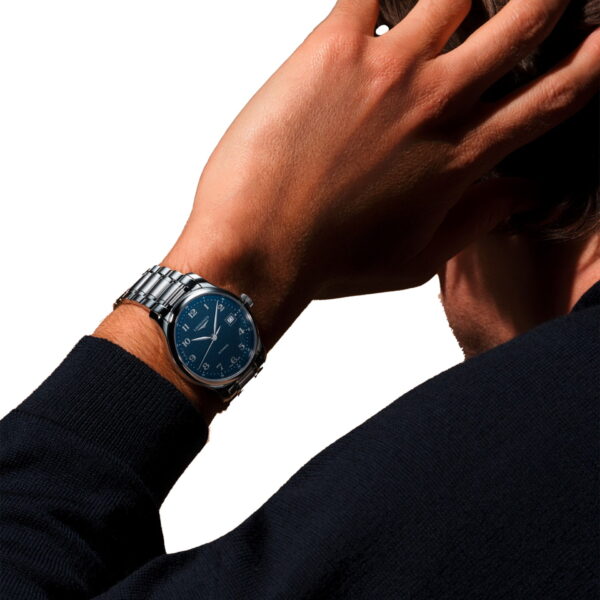 Eine Person in einem dunklen Pullover führt die Hand an den Kopf und präsentiert dabei die silberne Armbanduhr der Longines Master Collection mit blauem Zifferblatt und weißen Ziffern auf weißem Hintergrund.