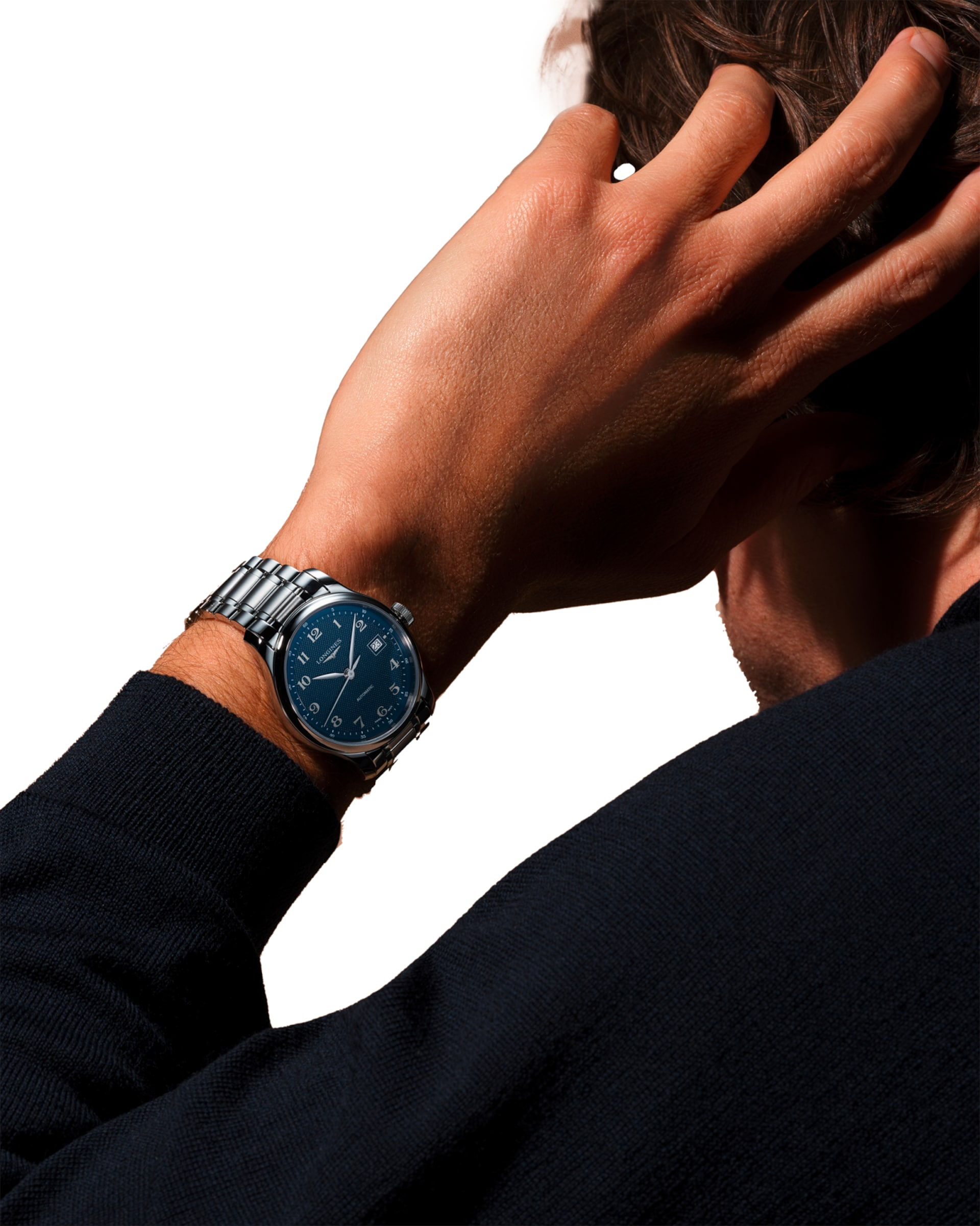 Eine Person in einem dunklen Pullover führt die Hand an den Kopf und präsentiert dabei die silberne Armbanduhr der Longines Master Collection mit blauem Zifferblatt und weißen Ziffern auf weißem Hintergrund.