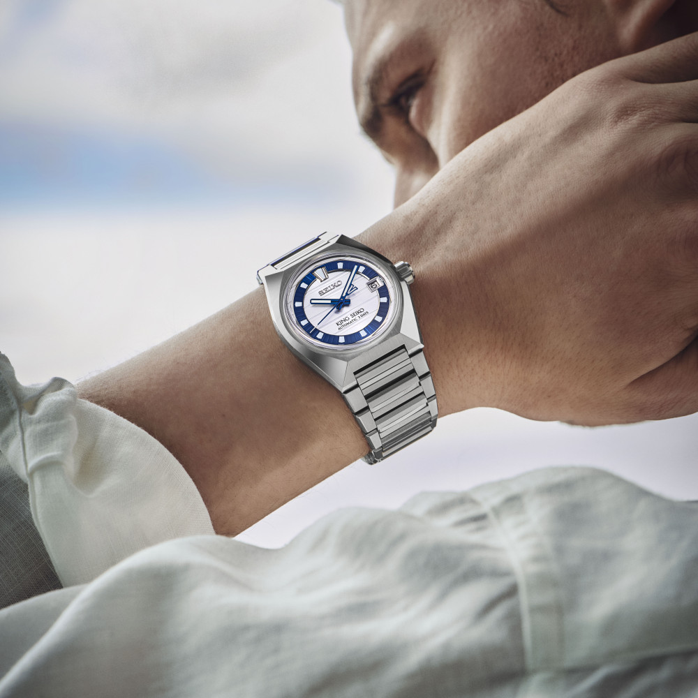 Eine Person in einem hellen Langarmhemd präsentiert die Seiko King Seiko Vanac Limited Edition – eine silberne Armbanduhr mit blau-weißem Zifferblatt –, indem sie ihr Handgelenk vor dem Hintergrund eines verschwommenen Himmels nahe an ihr Gesicht hält.