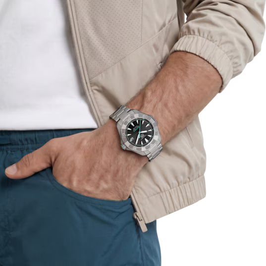 Eine Person, die eine beige Jacke und eine blaue Hose trägt, hat die linke Hand in der Tasche und präsentiert dabei die TAG Heuer Aquaracer Professional 200 Solargraph Armbanduhr mit schwarz-grünem Zifferblatt.
