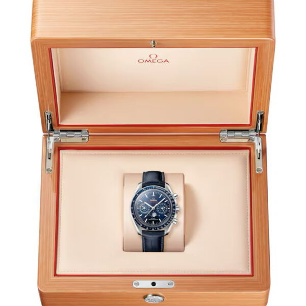 Eine OMEGA Speedmaster Moonwatch mit schwarzem Zifferblatt wird in einer hellen Holzschatulle mit beigem Innenfutter und roten Zierleisten präsentiert, auf deren Deckel das Omega-Logo zu sehen ist.