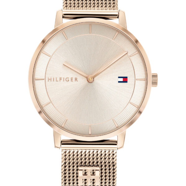 Eine Tommy Hilfiger Tea-Uhr mit einem goldenen Netzarmband, dessen Verschluss mit zwei Sternsymbolen verziert ist, einem minimalistischen Zifferblatt mit schmalen Stundenindizes, dem Schriftzug „HILFIGER“ und einem kleinen Logo in Form einer rot-weiß-blauen Flagge.