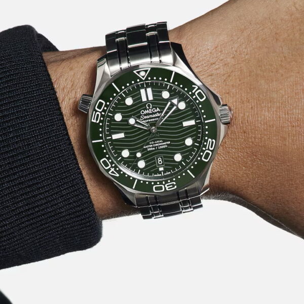 Eine Nahaufnahme einer Person, die eine OMEGA Seamaster Diver 300M mit grünem Zifferblatt und silbernem Edelstahlarmband am Handgelenk trägt; die Uhr zeigt 10:10 Uhr an.