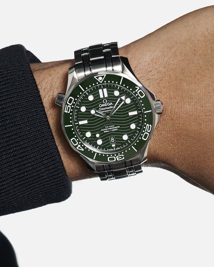 Eine Nahaufnahme einer Person, die eine OMEGA Seamaster Diver 300M mit grünem Zifferblatt und silbernem Edelstahlarmband am Handgelenk trägt; die Uhr zeigt 10:10 Uhr an.