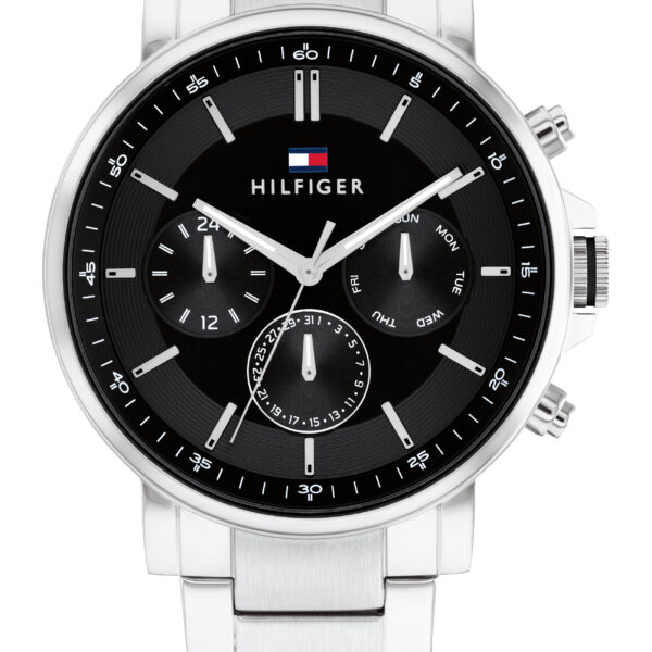 Die Tommy Hilfiger Tyson ist eine silberfarbene Metallarmbanduhr mit schwarzem Zifferblatt, drei Hilfszifferblättern und dem charakteristischen Hilfiger-Logo oben. Sie verfügt über ein Edelstahlarmband und mehrere Einstellknöpfe an der Seite.