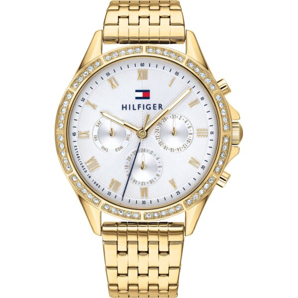 Die Tommy Hilfiger Ari Armbanduhr verfügt über ein goldenes Metallgliederarmband, ein weißes Zifferblatt mit römischen Ziffern als Stundenmarkierungen, drei Hilfszifferblätter sowie funkelnde Kristalle, die die Lünette verzieren.