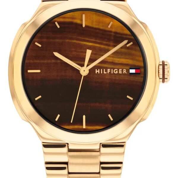 Tommy Hilfiger Georgia Armbanduhr mit einem goldenen Metallgliederarmband, einem braunen Zifferblatt mit Holzoptik, minimalistischem Design, goldenen Zeigern und dem „HILFIGER“-Logo, das durch eine kleine rot-weiß-blaue Flagge auf der rechten Seite akzentuiert wird.