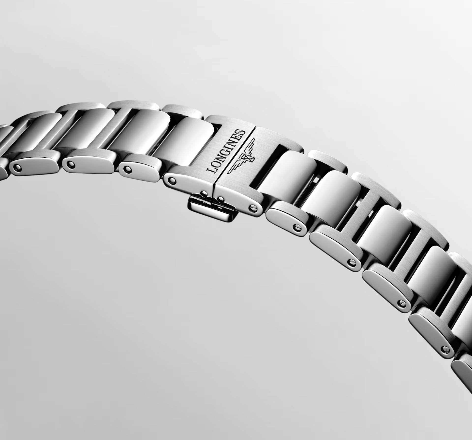 Eine Nahaufnahme des silbernen Metallarmbands der Longines Conquest, auf dessen einem Glied das „LONGINES“-Logo und das Symbol der geflügelten Sanduhr eingraviert sind, vor einem hellgrauen Hintergrund.